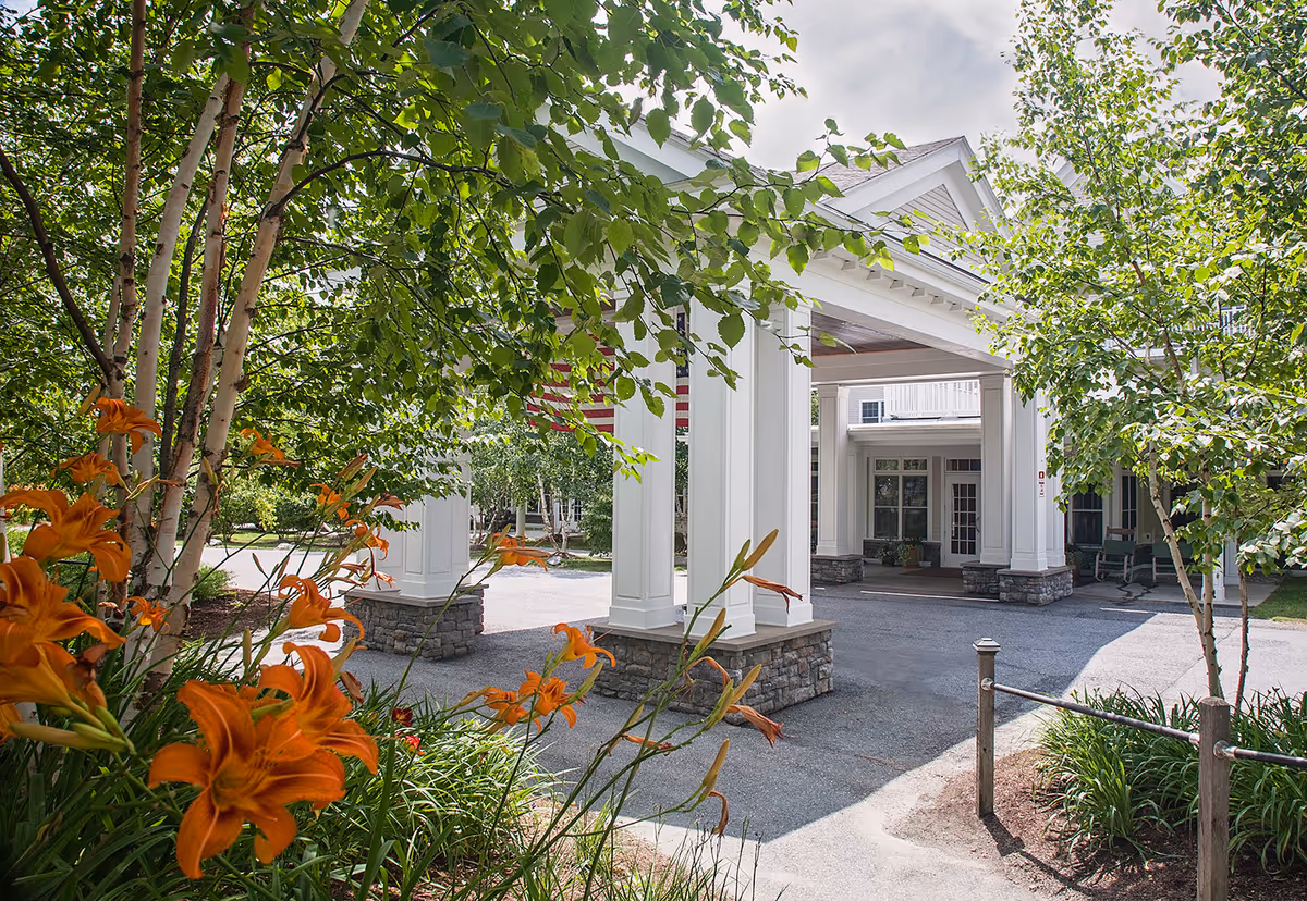 Entrance area of a senior living facility with white columns and a covered driveway surrounded by green trees and orange flowers in the foreground.