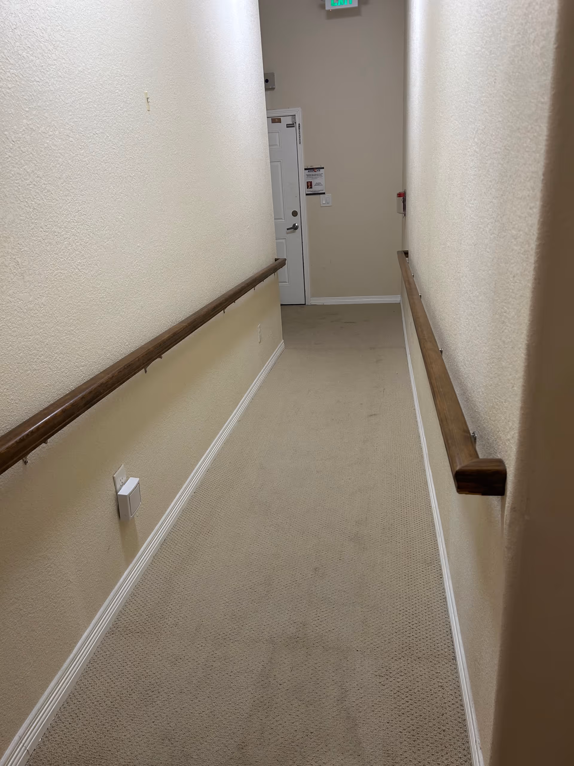 Long beige corridor with carpet, wood handrails, and a door at the end.