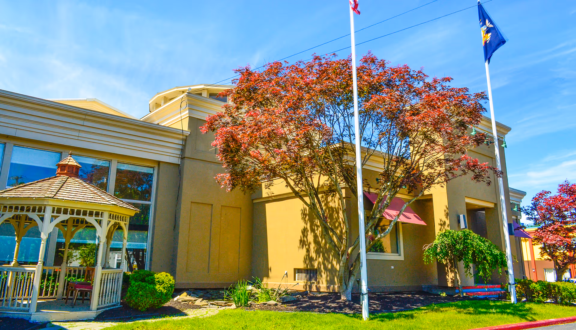 Front exterior of a beige senior living building with a gazebo, flagpoles, and red-leaved trees on a sunny day.