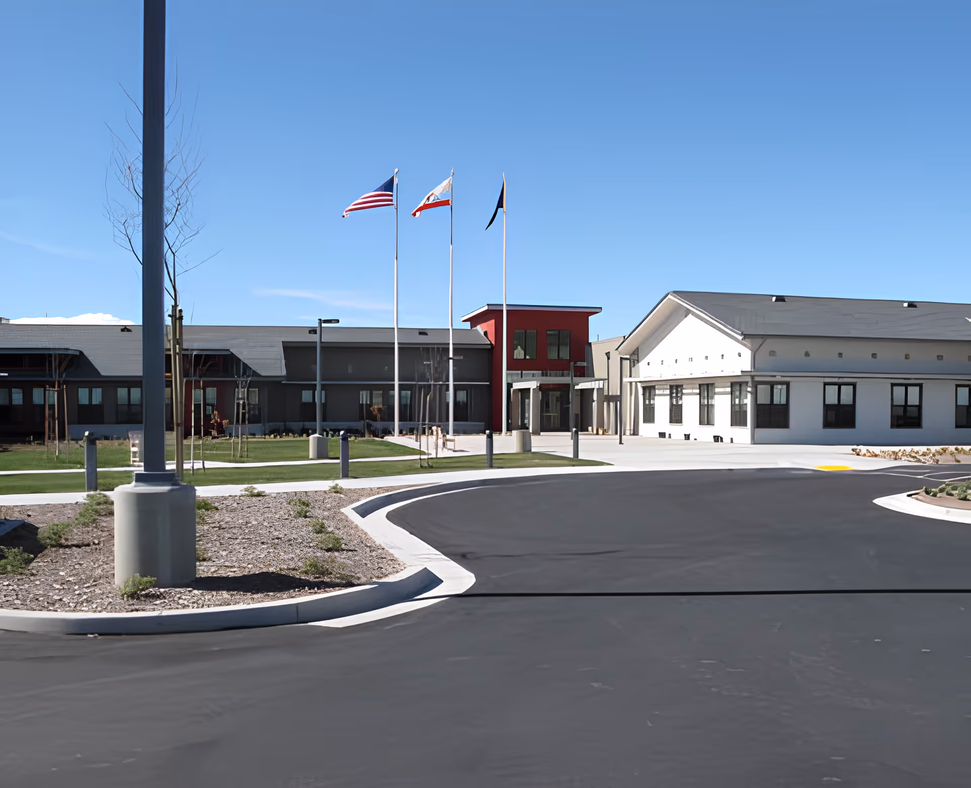 Exterior view of the William J. Pete Knight Veterans Home-Lancaster building with a clear blue sky, three flagpoles flying the American flag, California state flag, and another flag, a paved driveway, and landscaped areas with grass and small plants.