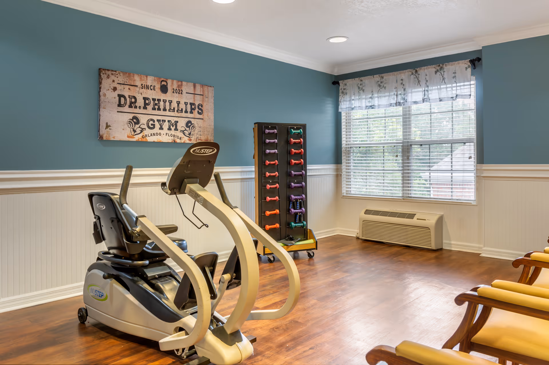 A small fitness room with an exercise bike, dumbbell rack, chairs, and a window.
