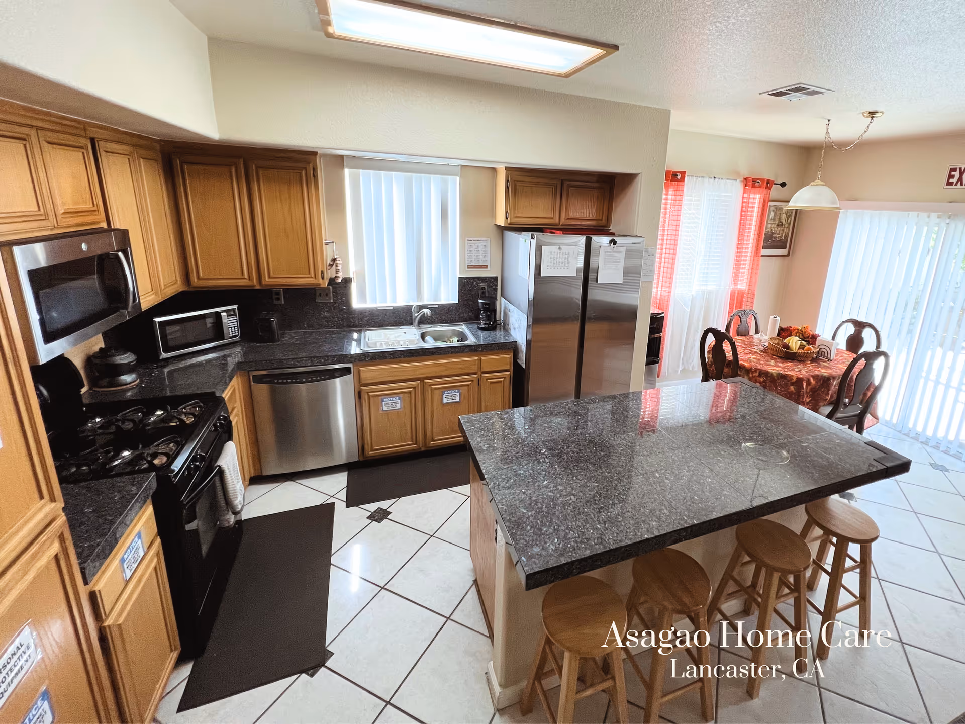 Bright kitchen with a large granite island and stools, wooden cabinets, stainless appliances, and a dining table by sliding glass doors.