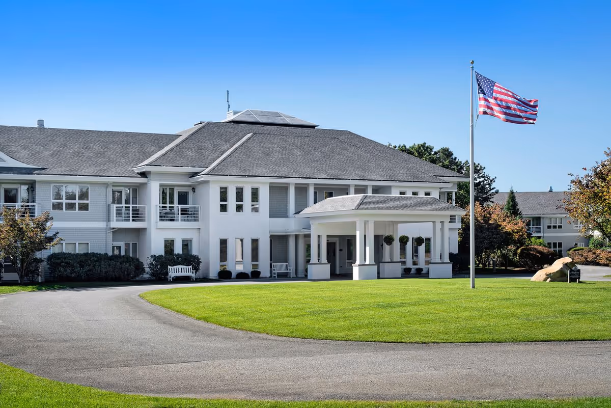 White senior living facility with a covered entrance, manicured lawn, and an American flag.