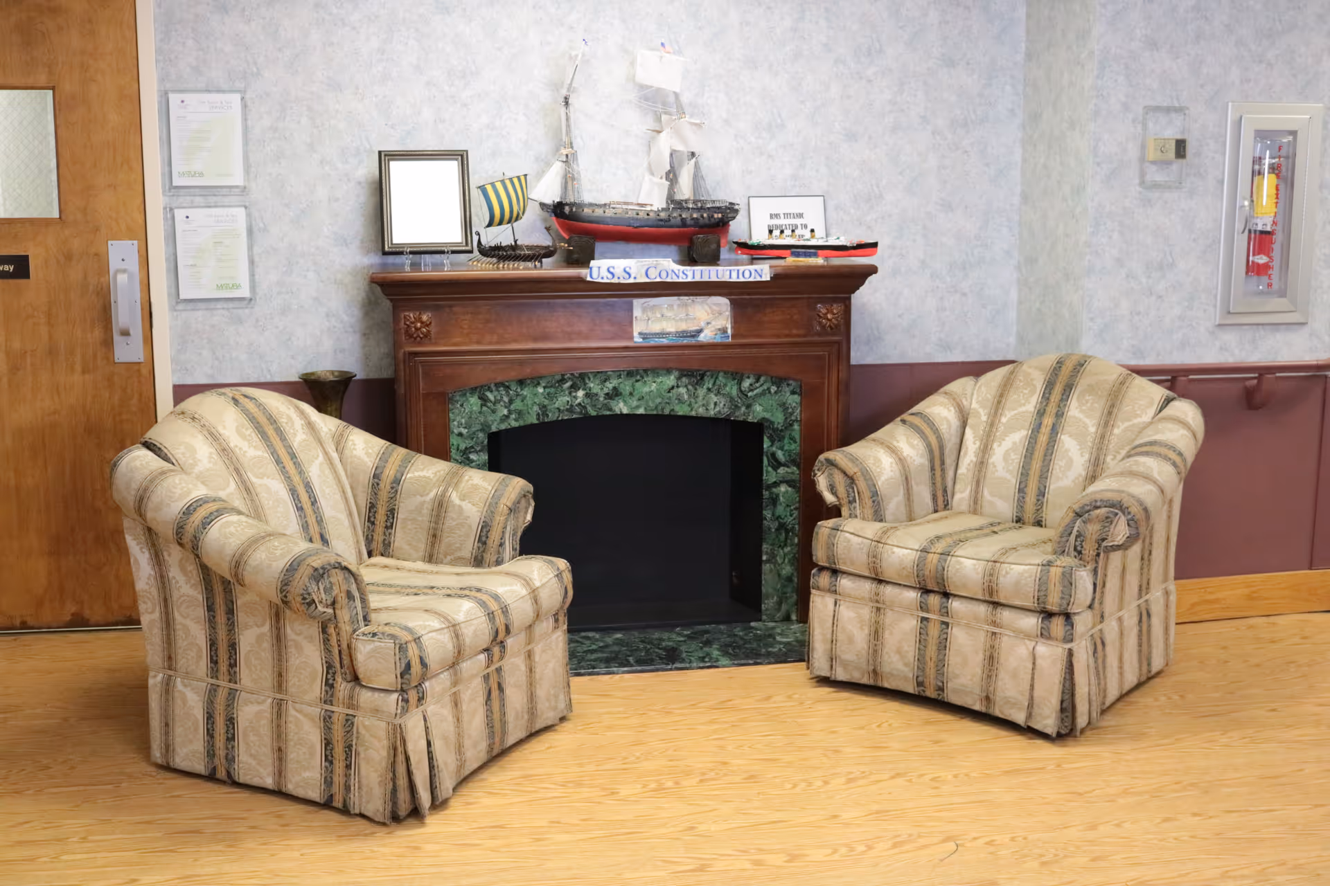 Two patterned armchairs facing each other in front of a decorative fireplace with a green marble surround. On the mantel are model ships, a framed picture, and a sign that reads 'U.S.S. Constitution'. The room has light-colored walls and a wooden floor.