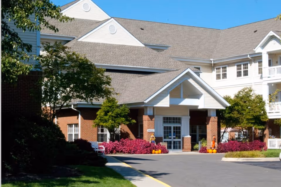 Front entrance of a light-colored senior living building with a covered porte-cochère, landscaping, and pumpkins by the doorway.