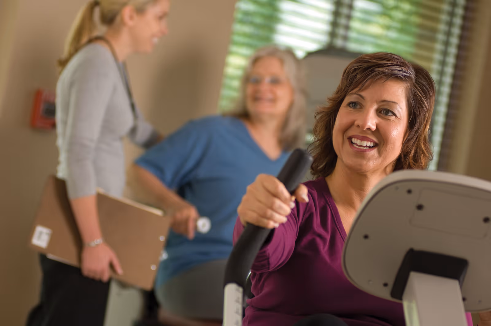 A woman in a purple top is using an exercise machine, smiling. In the background, a woman in blue scrubs is seated and smiling, while another woman holding a clipboard stands nearby, engaging with them.