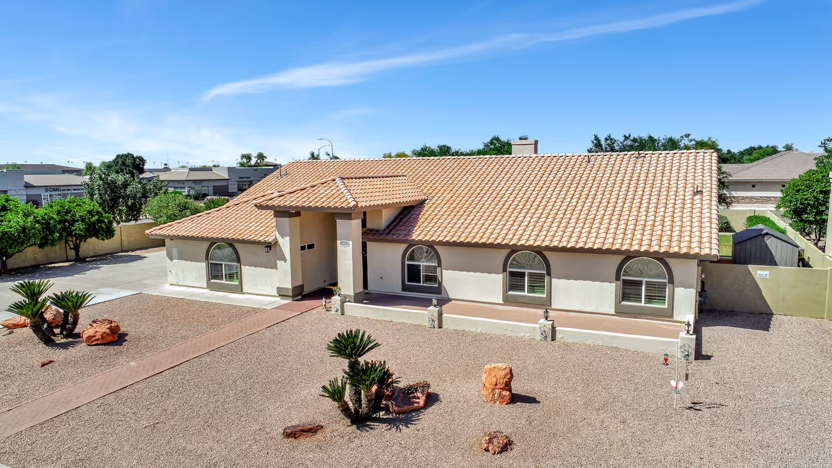 Single-story stucco building with a tiled roof and arched windows set behind a gravel, desert-style front yard under a clear blue sky.