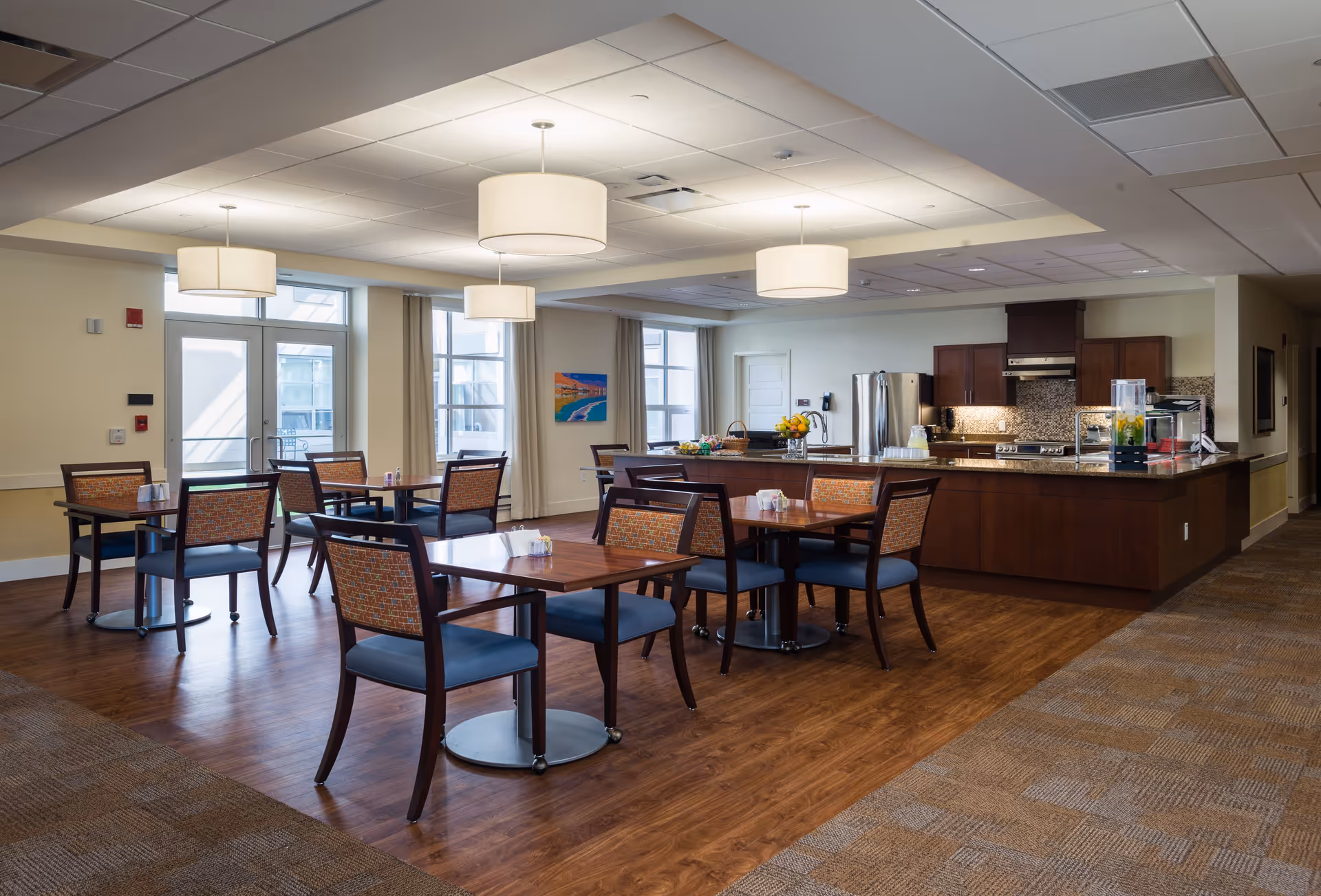 Dining area with several wooden tables and chairs, pendant lights, and a kitchen/service counter in the background.