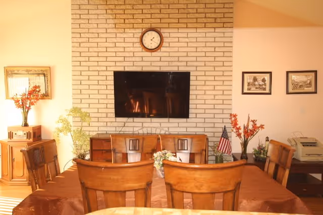 A cozy dining area with a wooden table covered with a brown tablecloth and surrounded by six wooden chairs. On the table, there is a small floral centerpiece. Behind the table, a white brick wall features a mounted flat-screen TV and a round clock above it. To the left, there is a wooden cabinet with a vase of orange flowers and a framed mirror above it. To the right, there are two framed pictures on the wall, a small American flag, and additional floral arrangements on furniture pieces.