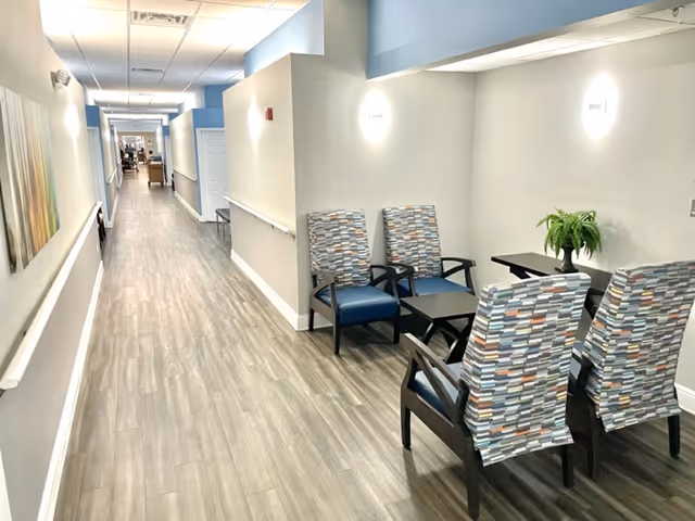 Long, well-lit hallway in a senior living facility with seating and a plant.