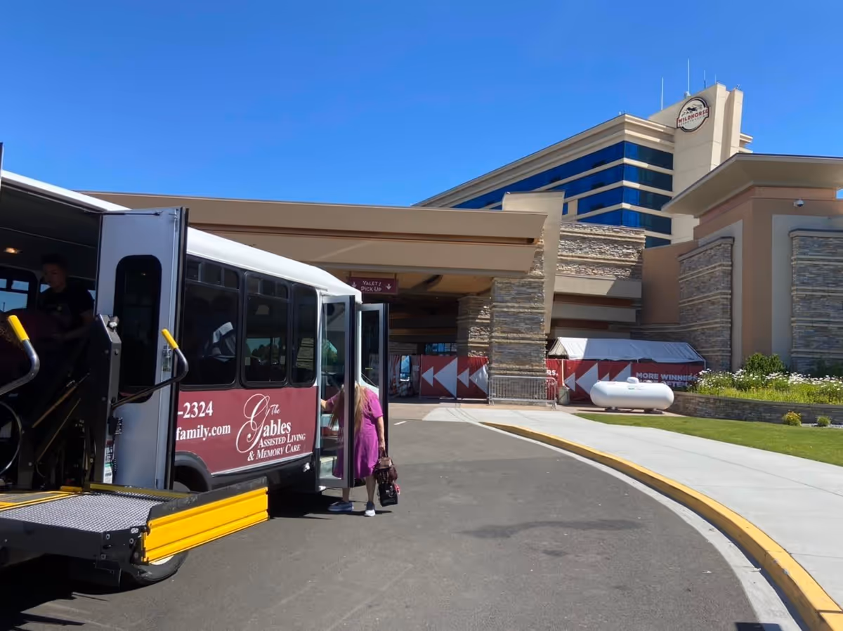 A shuttle bus with 'The Gables Assisted Living & Memory Care' written on its side is parked outside a large building under a clear blue sky. A person wearing a purple dress is boarding the bus. The building has stone and beige exterior walls with a covered entrance and a sign indicating valet and pick up. There is a tall building in the background with reflective blue windows.