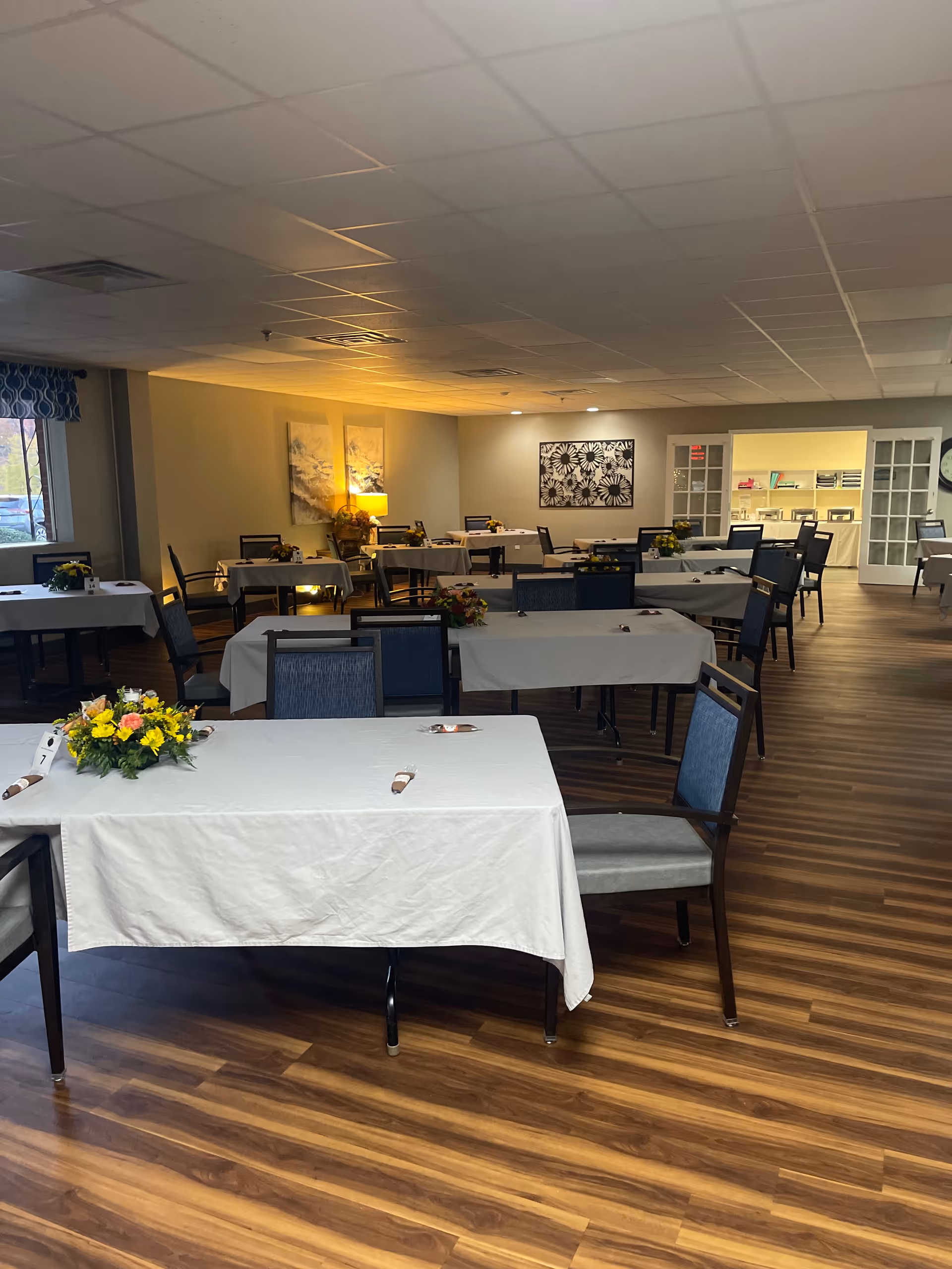 Empty dining room with white tablecloths, flowers, and multiple tables in a senior living facility.