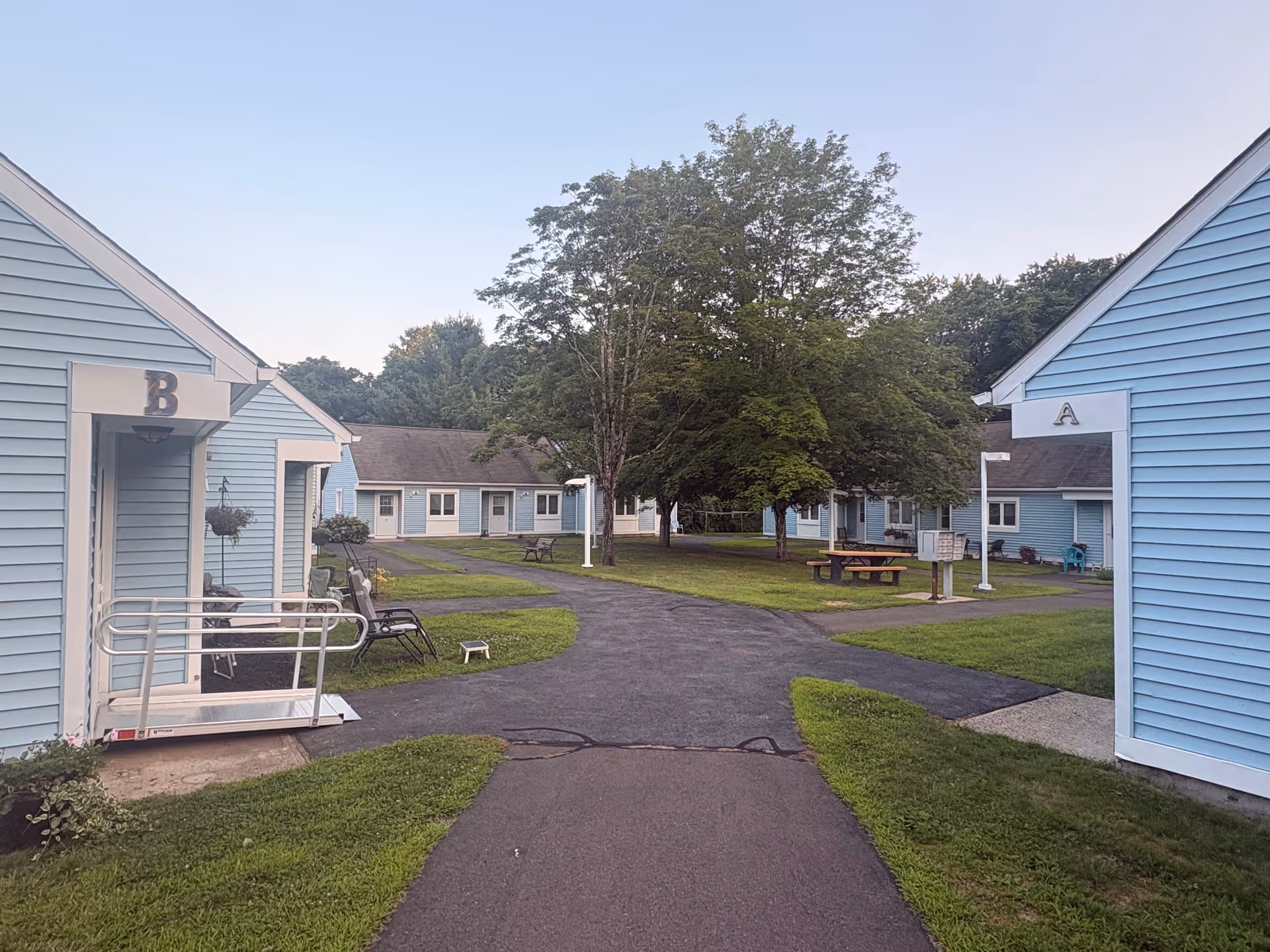 Blue single-story apartment-style buildings surround a grassy courtyard with trees and benches.