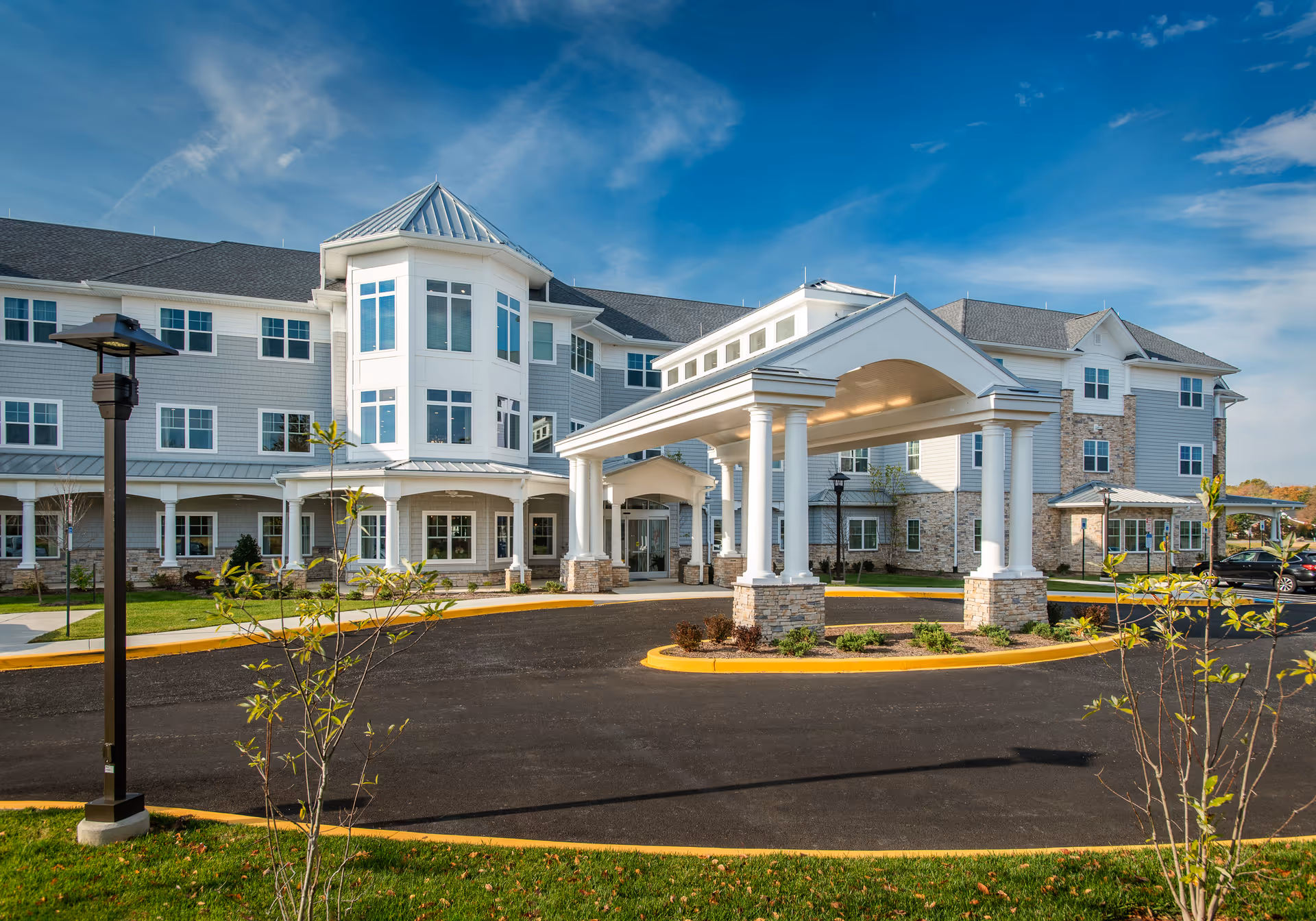 Exterior view of The Lodge at Historic Lewes, a large multi-story senior living facility with a covered entrance, stone and siding facade, surrounded by a driveway and landscaped greenery under a blue sky.