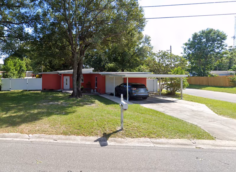 Red single-story facility with carport, driveway, mailbox, and large tree in front yard.