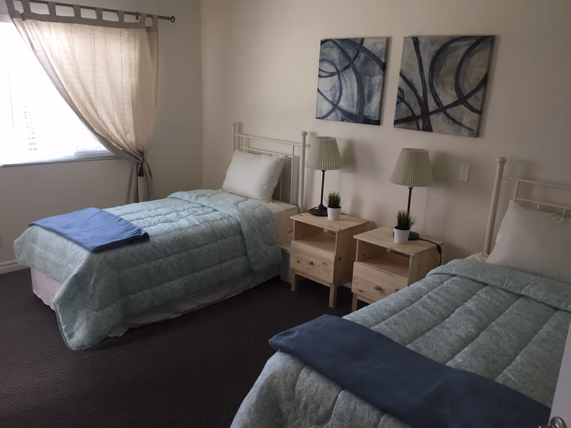Two single beds with matching bedding, nightstands, lamps, and abstract wall art in a bright bedroom.
