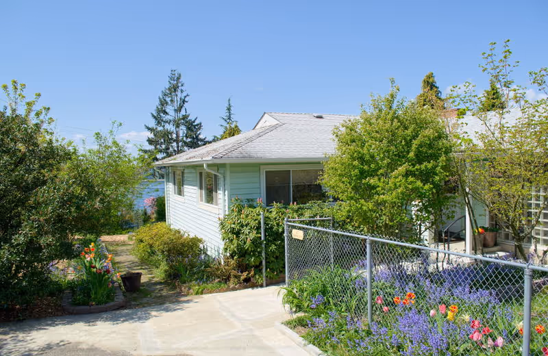 A single-story light-blue home with a garden, walkway, and chain-link fence.