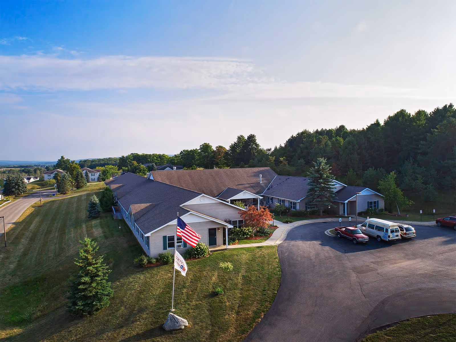 Aerial view of the American House Petoskey senior living facility showing a single-story building with a large parking area, surrounded by trees and greenery under a partly cloudy sky.