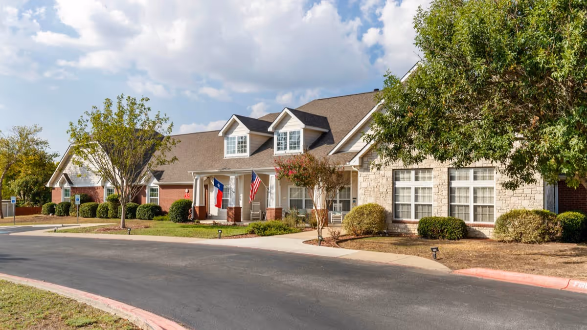 Front exterior of a single-story brick and stone senior living building with flags, landscaping, and a curved driveway.