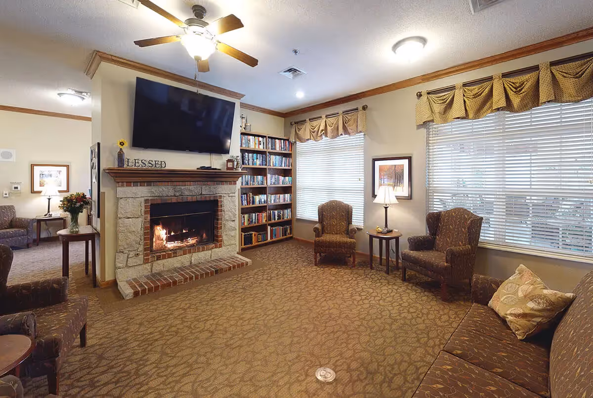 A cozy living room with a stone fireplace and a mounted flat-screen TV above it. There is a bookshelf filled with books next to the fireplace. The room has multiple upholstered chairs and a sofa with a decorative pillow. Two windows with blinds and valances let in natural light, and there are table lamps providing additional lighting. The carpet has a subtle pattern, and the walls are painted a warm beige color.