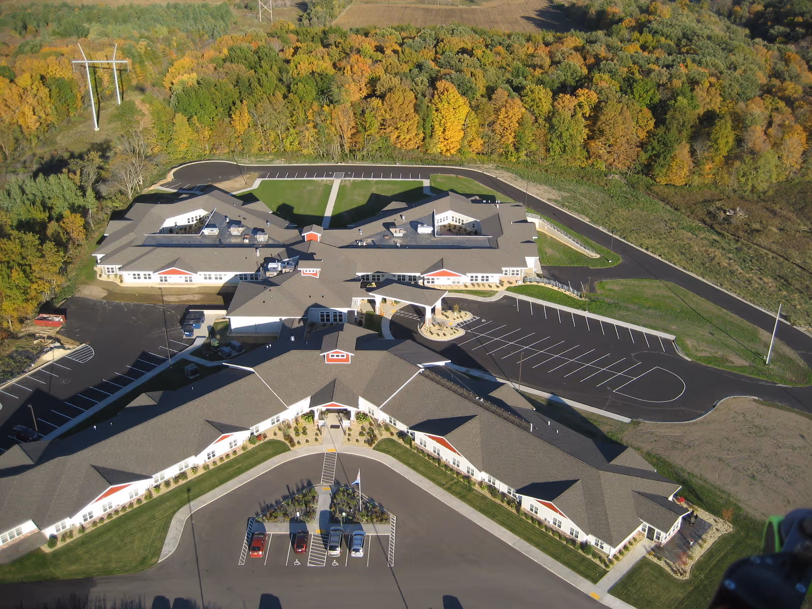 Aerial view of Valley Villas Assisted Living facility showing a large, single-story building with multiple wings, surrounded by parking lots and green lawns, with a backdrop of dense trees displaying autumn colors.