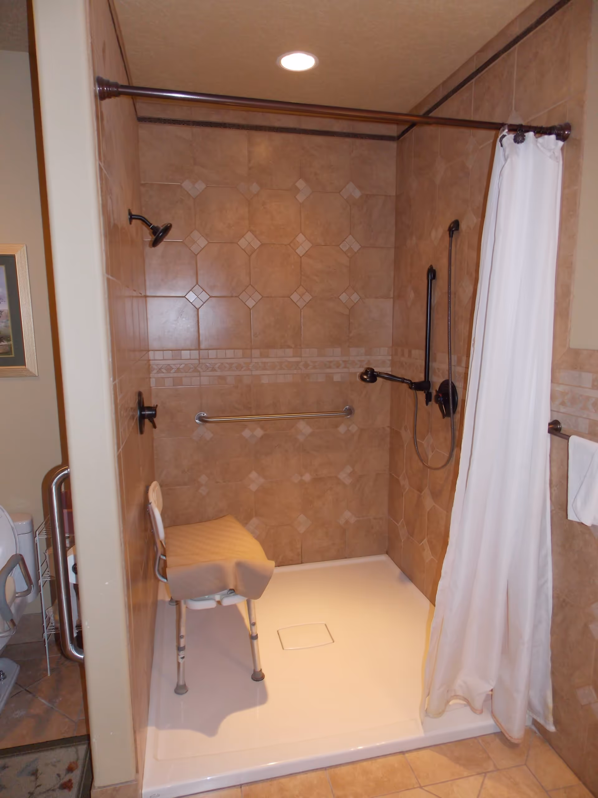 A tiled walk-in shower with a white shower curtain partially drawn to the side. Inside the shower is a white shower chair with a beige towel draped over it. The shower has a handheld showerhead and a fixed showerhead, along with a horizontal grab bar on the back wall. The tiles are a light brown color with a decorative pattern. Part of a toilet and a grab bar are visible to the left outside the shower.
