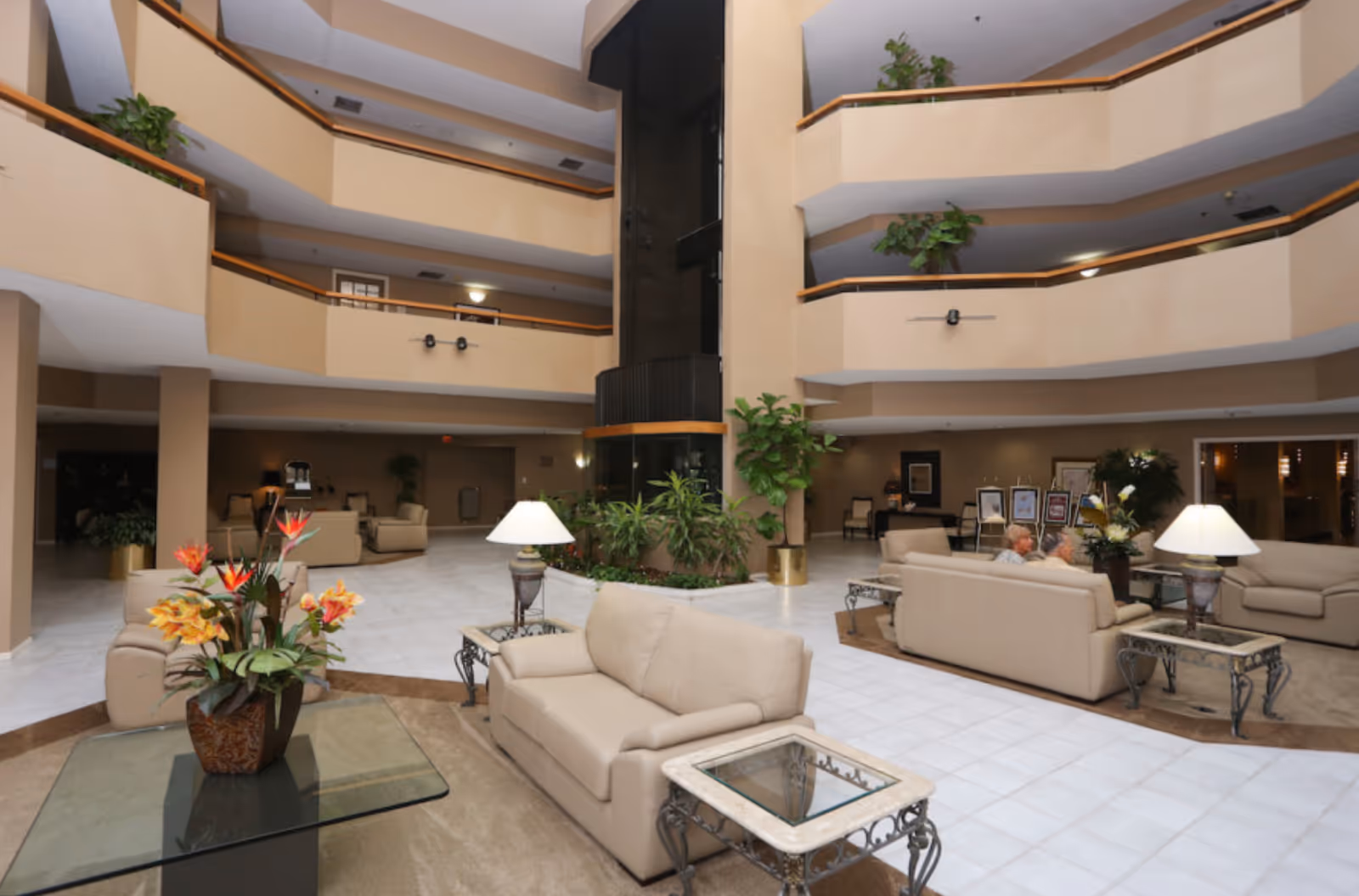 Spacious multi-level atrium lounge with beige sofas, side tables, plants, and a central glass elevator.