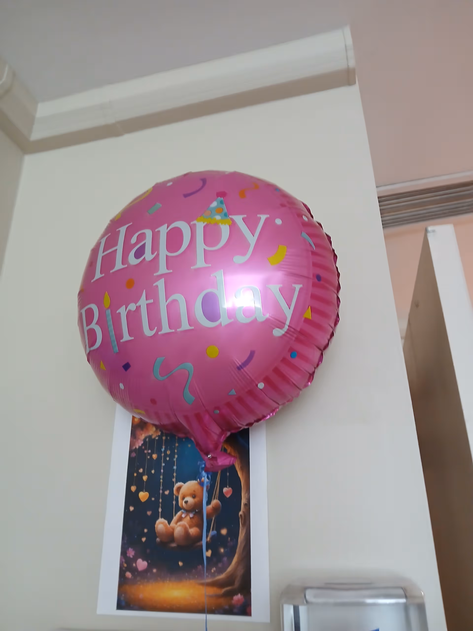Pink 'Happy Birthday' balloon attached to a wall in a facility hallway.
