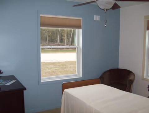 Blue-walled senior living bedroom with a bed, window, chair, and dresser.