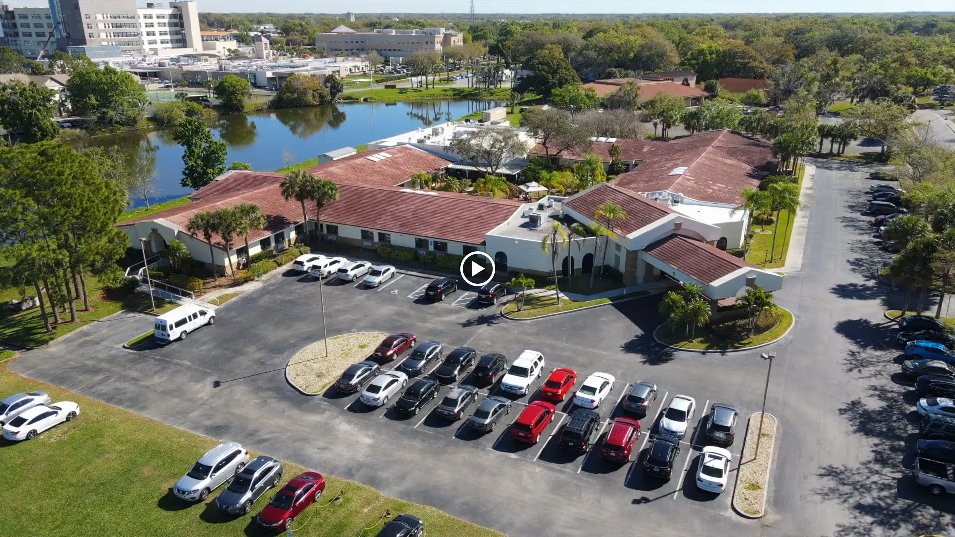 Aerial view of The Terrace of Kissimmee senior living facility showing a single-story building with a red-tiled roof surrounded by parking lots with multiple parked cars. The facility is adjacent to a pond with trees and greenery around it, and other buildings are visible in the background under a clear sky.