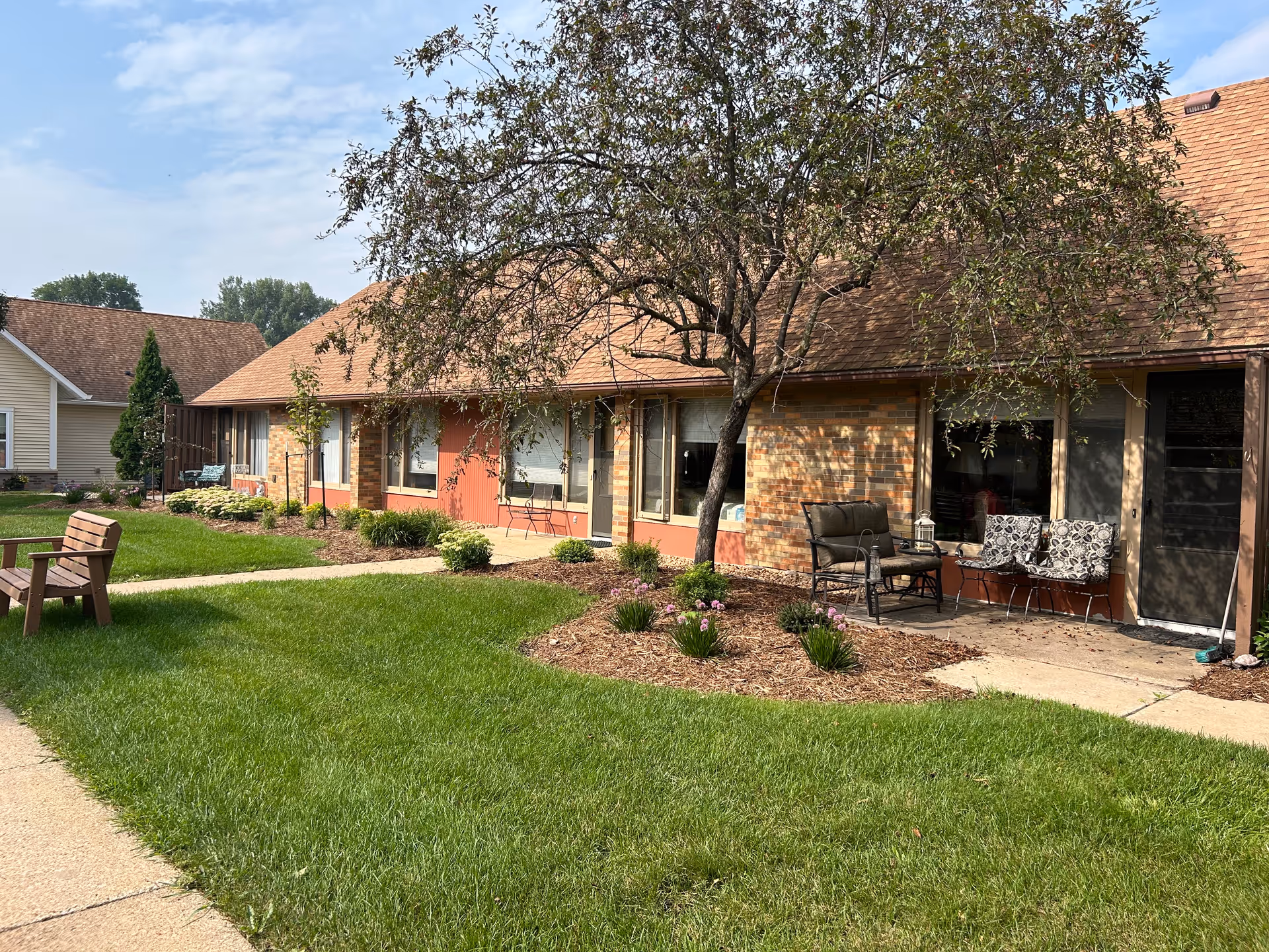 A single-story brick senior living building with a landscaped front yard and outdoor seating.