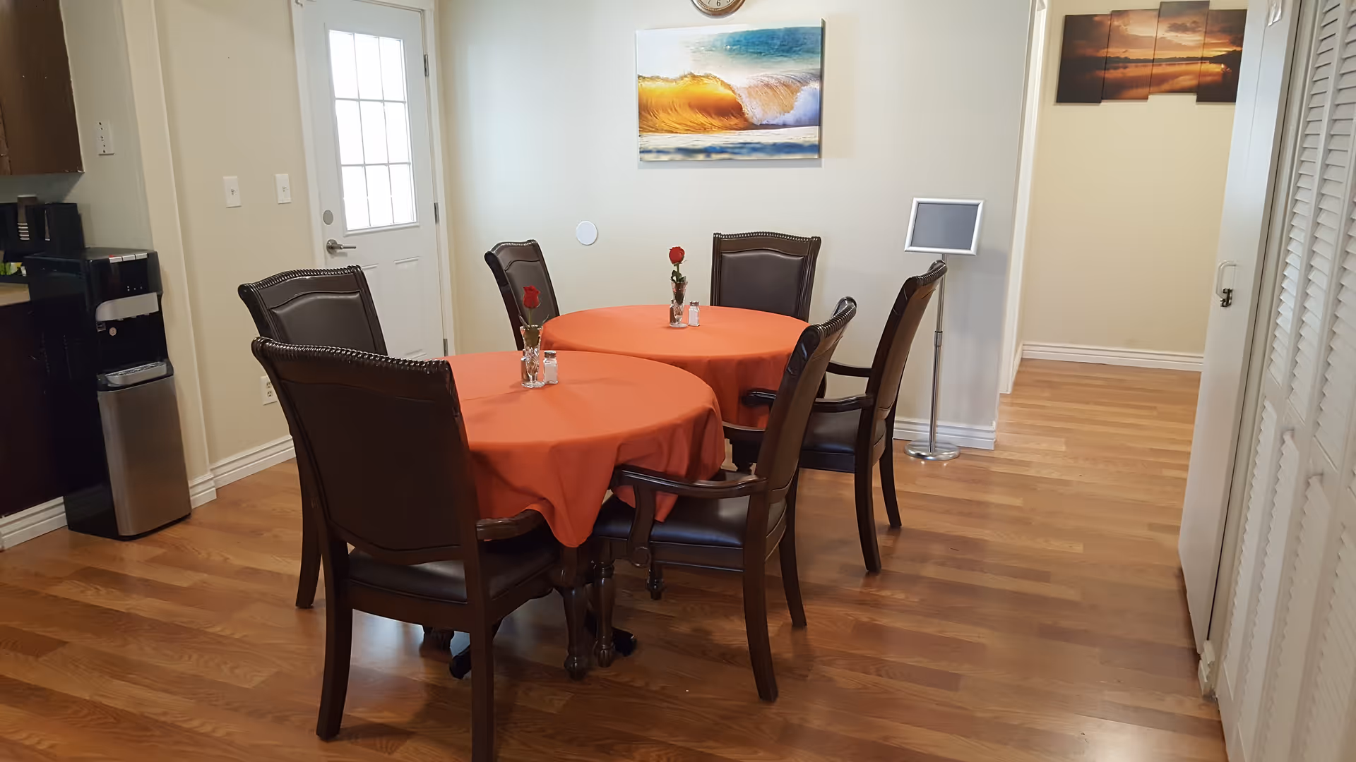 Two round tables with orange tablecloths and cushioned chairs in a bright dining room.