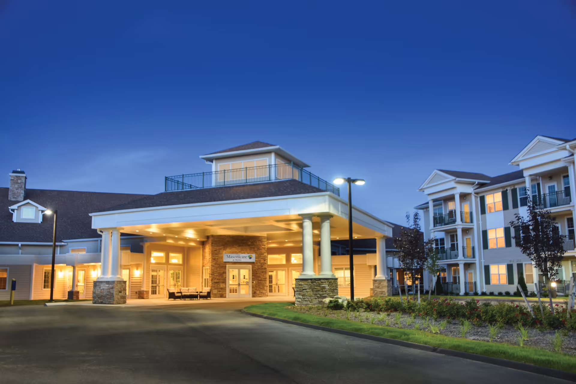 Exterior view of Masonicare At Mystic facility at dusk, showing the main entrance with a covered driveway supported by white columns and a well-lit building with multiple windows and balconies.