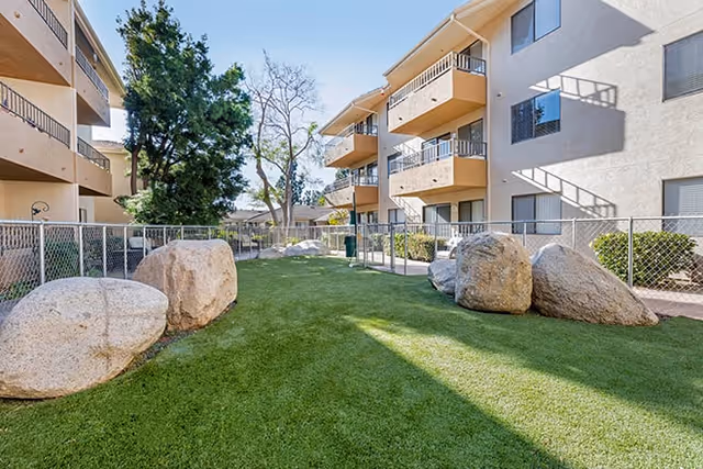 Outdoor courtyard area between two beige multi-story residential buildings with balconies, featuring a grassy lawn with large decorative rocks and a chain-link fence surrounding the space. Trees and shrubs are visible in the background under a clear blue sky.