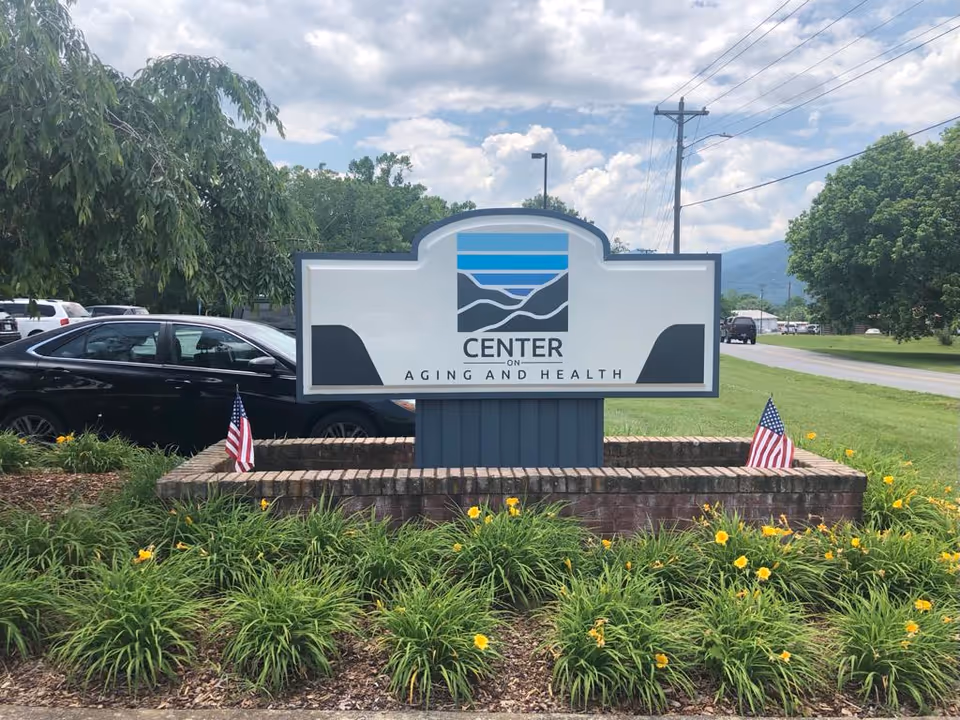 Outdoor sign reading "Center on Aging and Health" surrounded by flowerbeds and small American flags with parked cars and trees in the background.