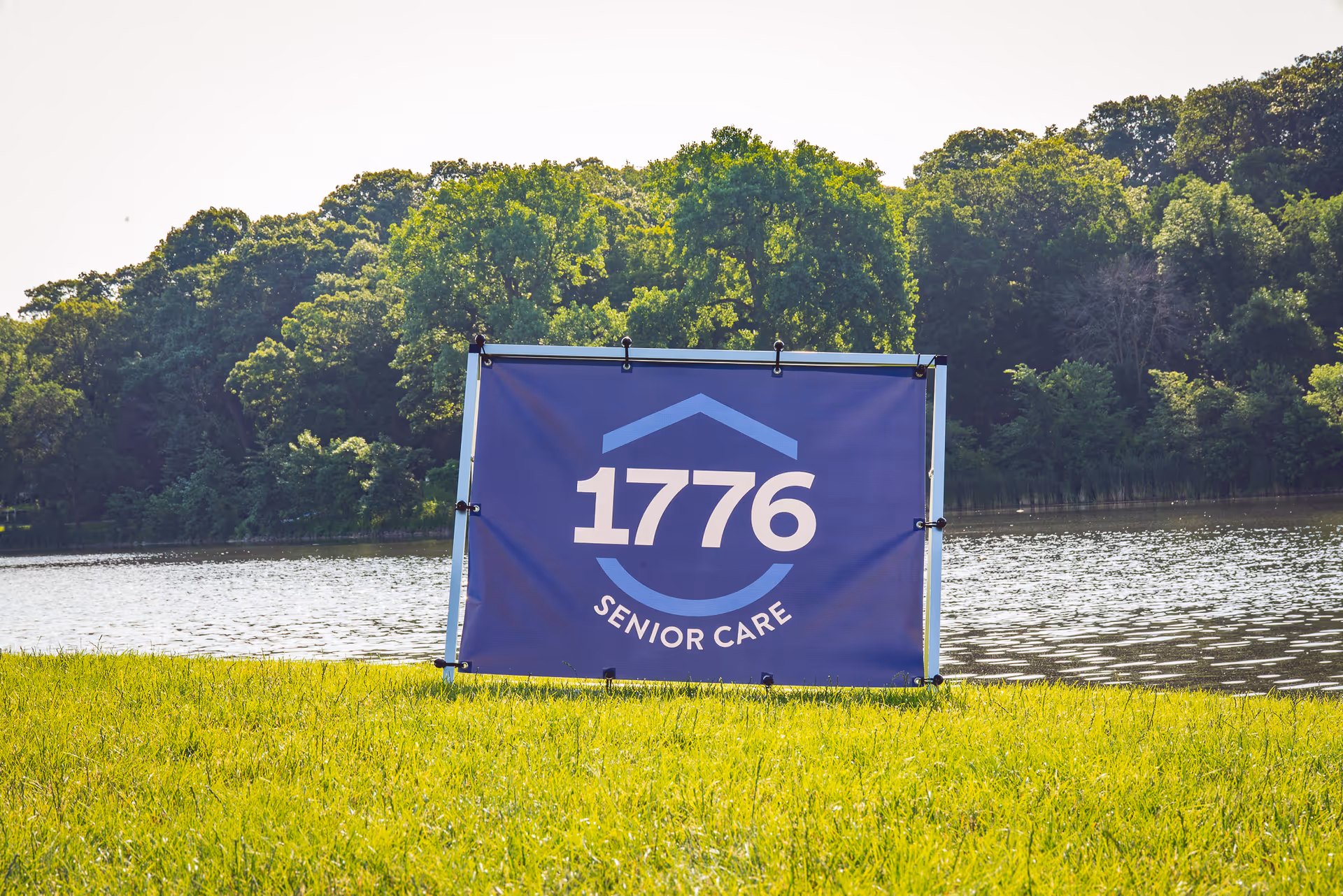 Blue sign reading 1776 Senior Care positioned on a grassy lakeside field.