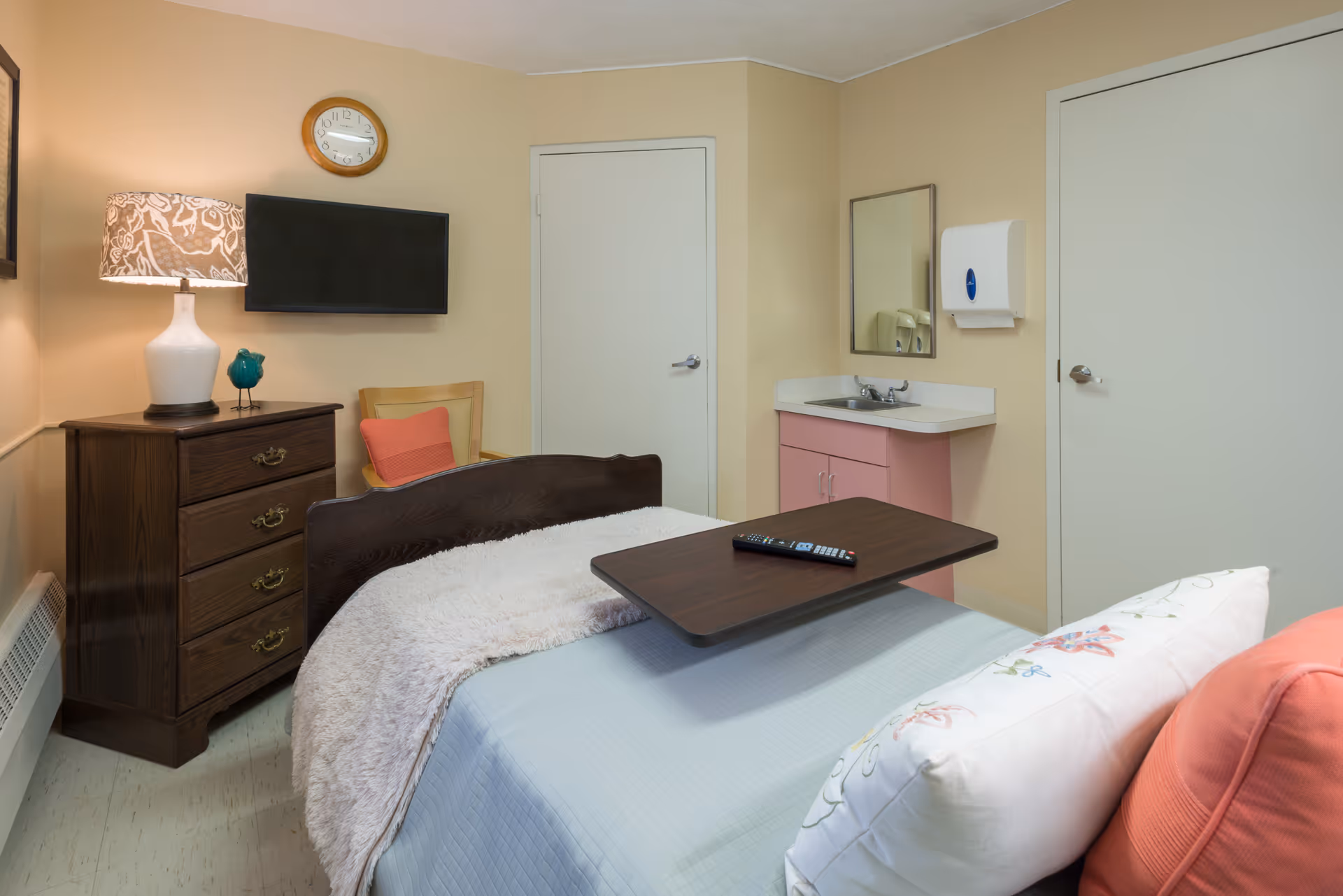 A small bedroom in a senior living facility featuring a bed with a light blue blanket and floral pillow, a dark wooden overbed table with a remote control on it, a wooden dresser with a white lamp and decorative bird, a wall-mounted flat-screen TV, a clock above the TV, a wooden chair with a coral cushion, a small sink with a mirror and paper towel dispenser, and two closed doors.