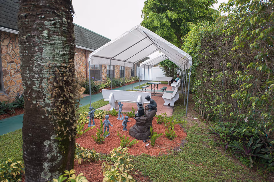 A covered outdoor seating area with white benches and tables beside a landscaped garden bed with small statues along the side of a building.