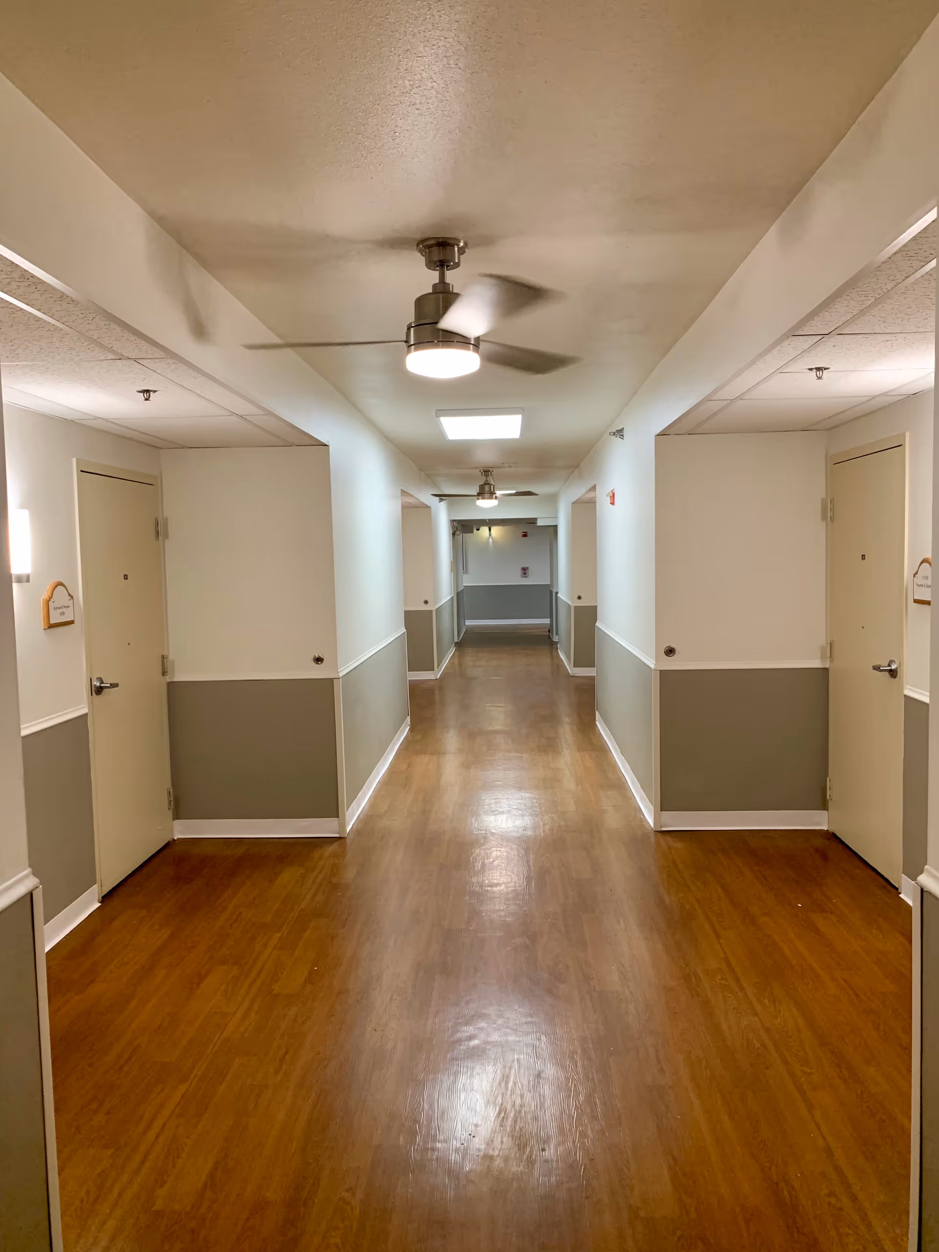 Long indoor corridor in a senior living facility with doors on both sides and wood flooring.