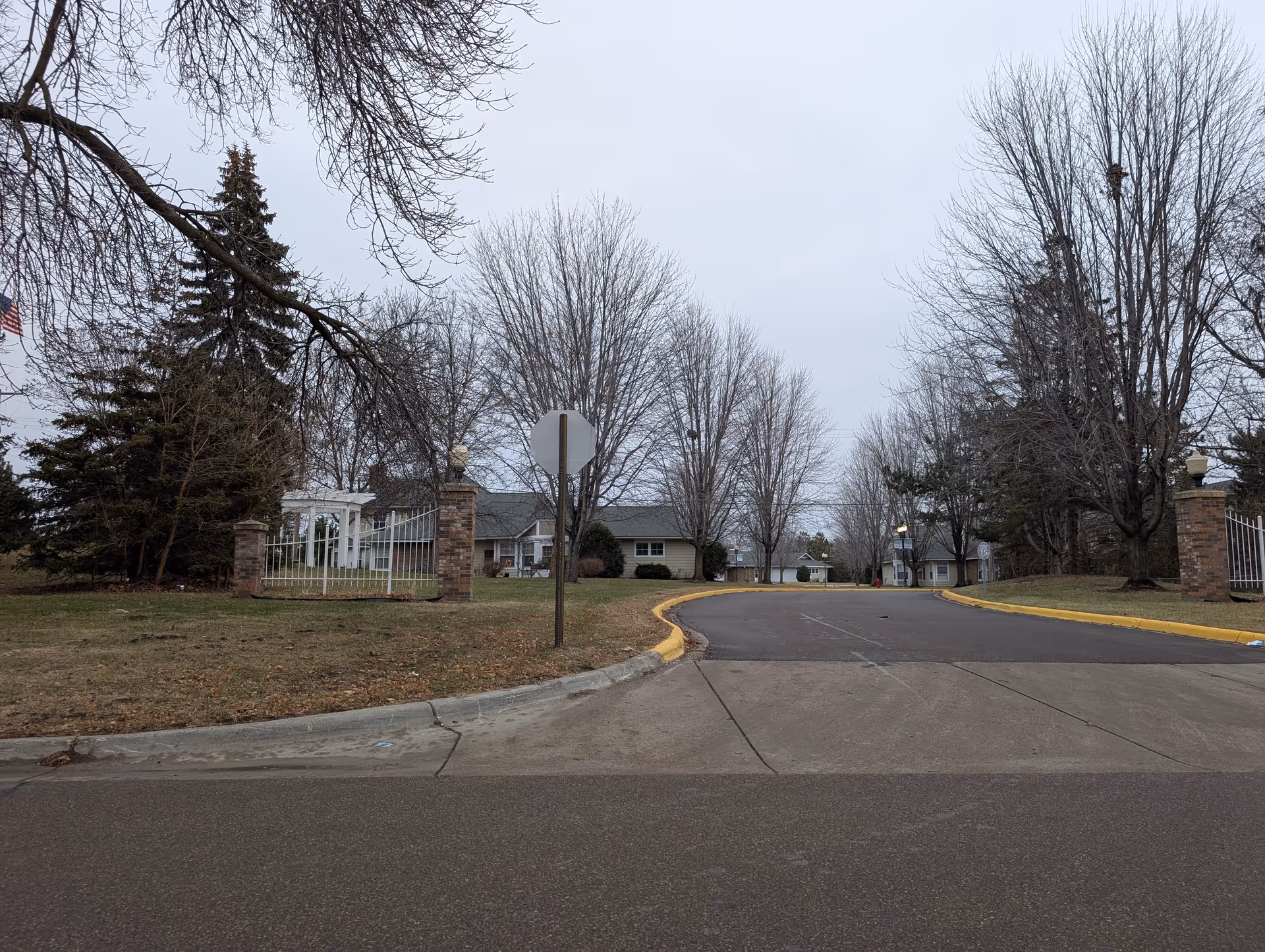 Entrance driveway to Waterford Manor with gated brick pillars, leafless trees, and single-story buildings under a cloudy sky.