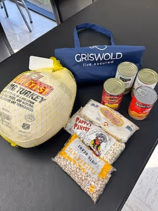 Assorted canned foods and a large poultry bag beside a Griswold tote on a dark table.