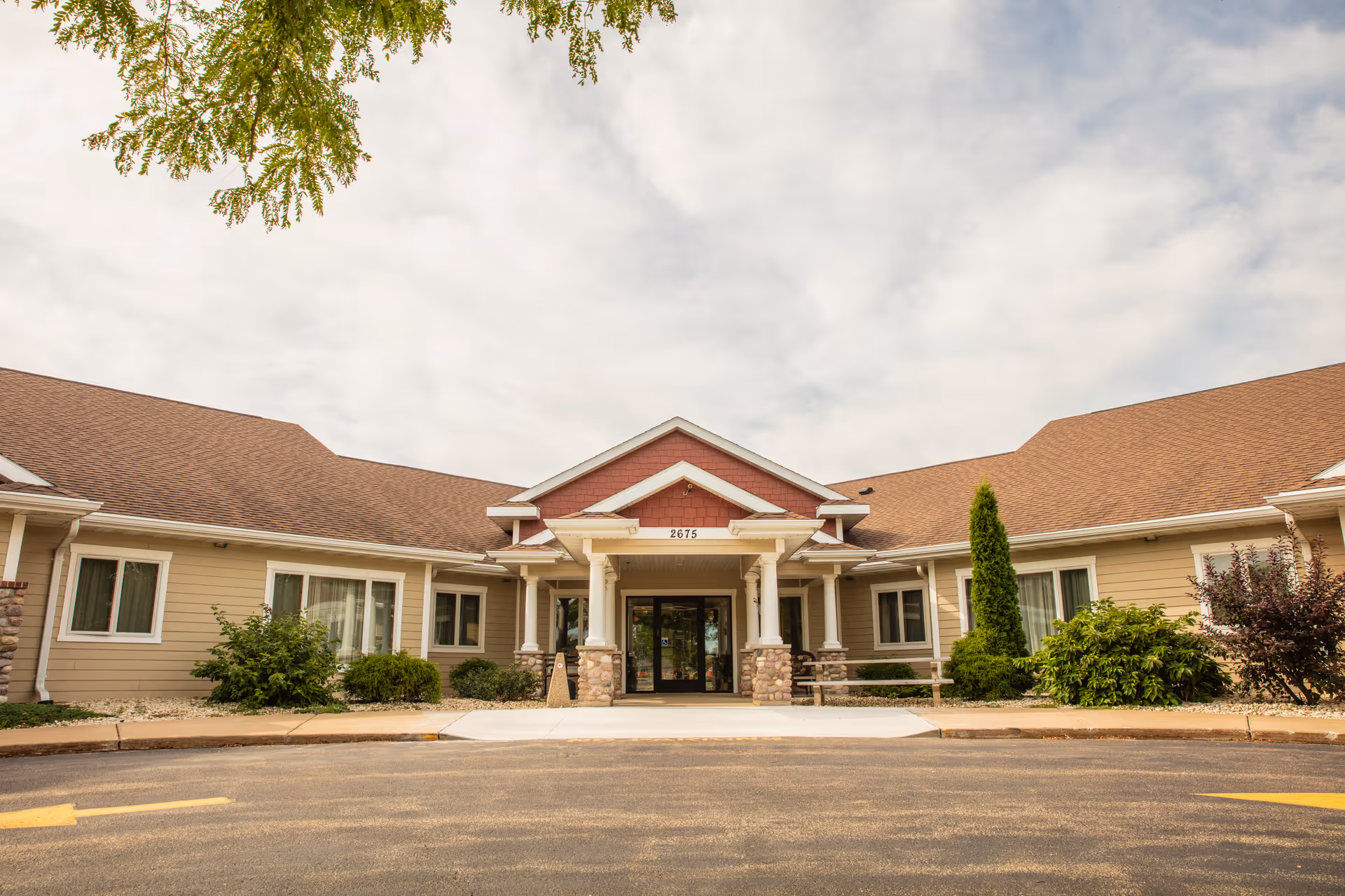 Front exterior view of Oakwood Manor, a single-story building with beige siding, a brown shingled roof, and a covered entrance supported by stone pillars. There are shrubs and small trees around the building and a paved driveway in front.