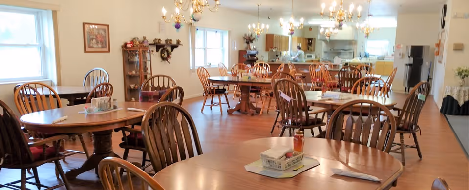 A spacious dining hall with round wooden tables and chairs, kitchen area in the background.