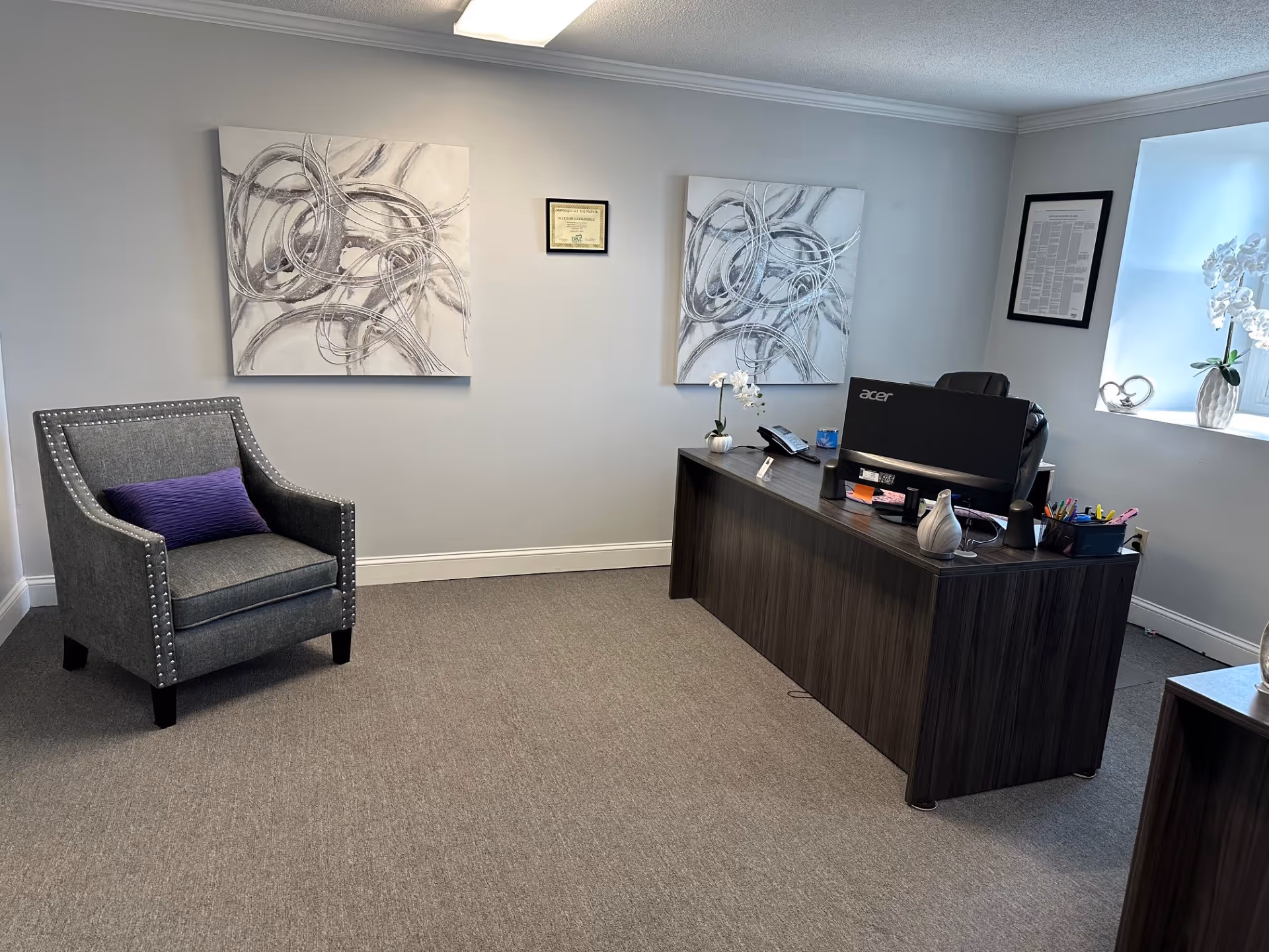 A modern office reception area with a dark wood desk, gray armchair, and abstract wall art.