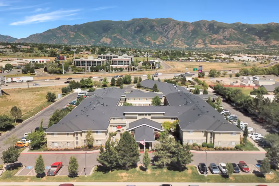 Aerial view of Chancellor Gardens senior living facility with a large building surrounded by parking lots, trees, and landscaping. In the background, there is a highway with cars and trucks, commercial buildings, and a mountain range under a clear blue sky.