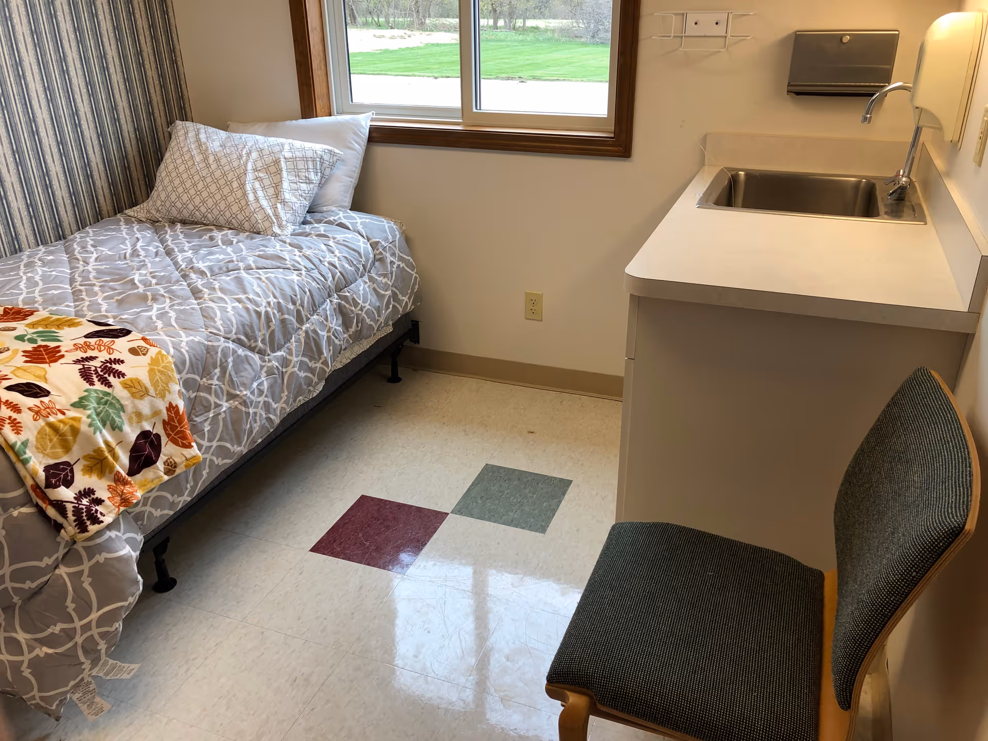 A small room with a single bed covered with a gray patterned comforter and a colorful blanket with leaf designs. There is a pillow on the bed. Next to the bed is a countertop with a built-in sink and a chair with a dark green cushion. A window above the bed shows a view of green grass outside.