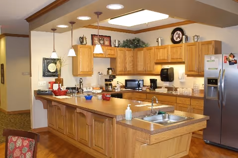 A spacious kitchen with wooden cabinets and a large island countertop featuring a sink and hand sanitizer. The kitchen is equipped with a stainless steel refrigerator, microwave, coffee maker, and various kitchen utensils. Pendant lights hang above the island, and decorative items are placed on top of the cabinets.