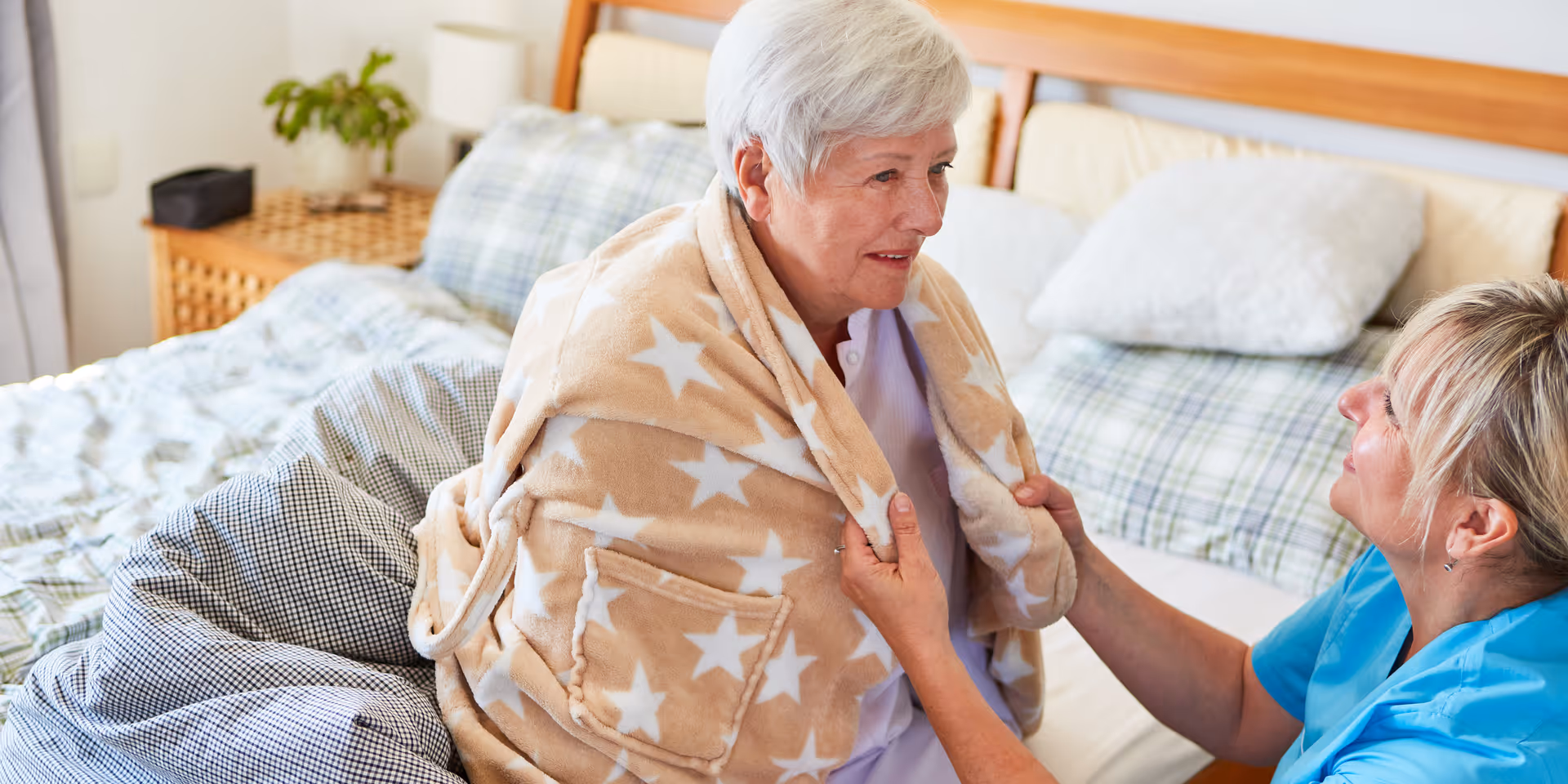 An elderly woman wrapped in a beige blanket with white stars sits on a bed while a caregiver in blue scrubs kneels beside her, holding the blanket and engaging in a warm conversation in a cozy bedroom setting.