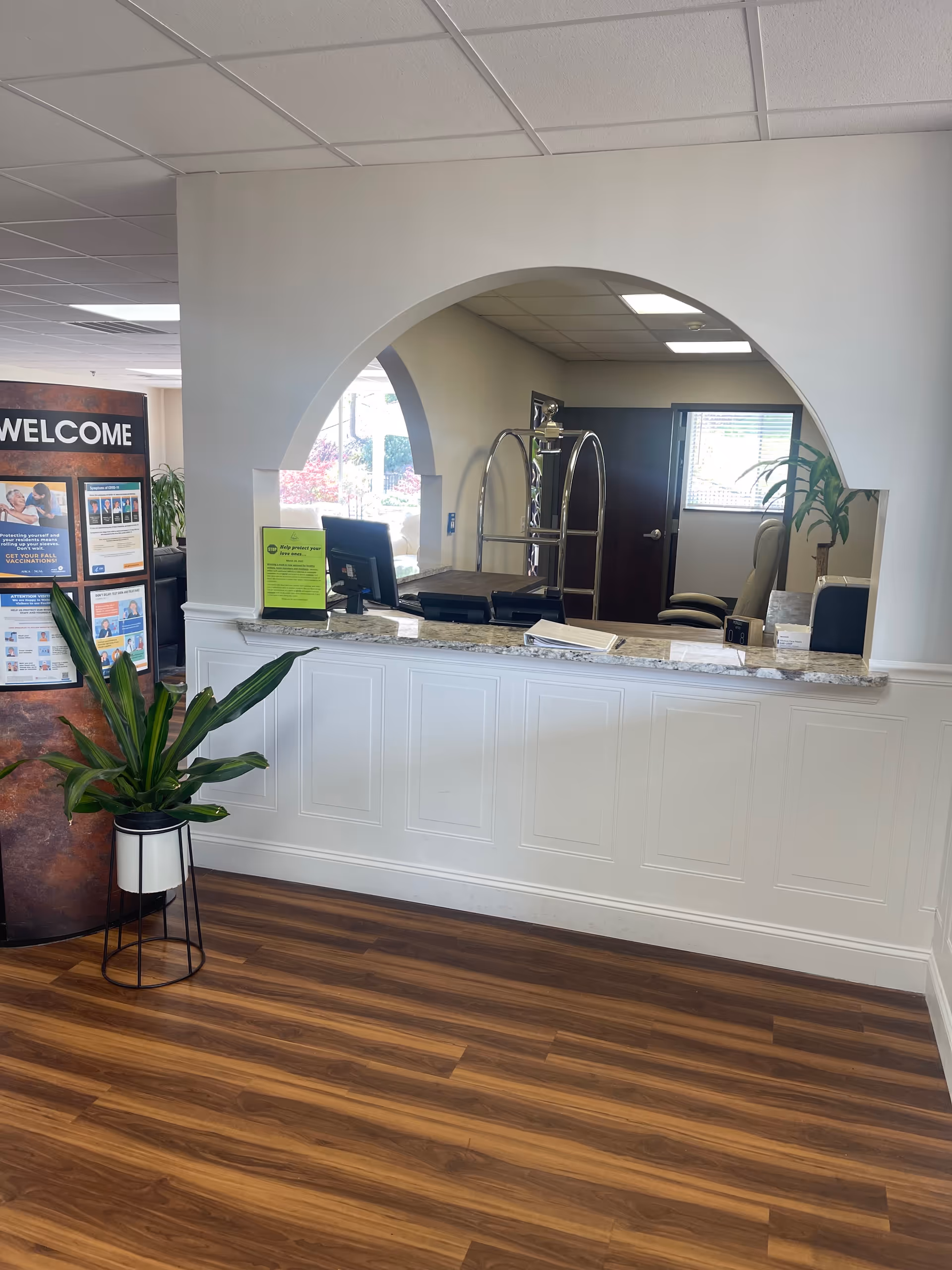 Reception desk with arched opening, wood floor, plants, and a welcome sign inside.