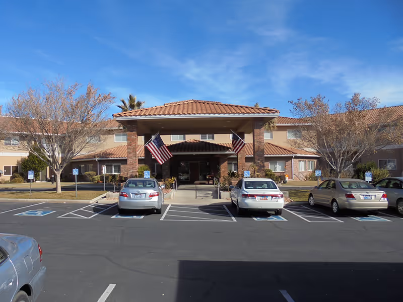 Front entrance of a two-story assisted living building with a covered porte-cochère, American flags, and parked cars in front.
